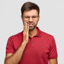 Photo of Caucasian man suffers painful toothache, has rotten tooth, needs to visit dentist, keeps eyes closed from pain, dressed in casual red t shirt, isolated over white background, touches cheek